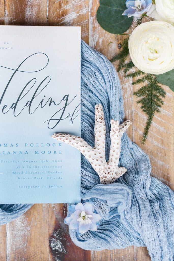 the wedding invitation with shells and blue flowers with greenery surrounding