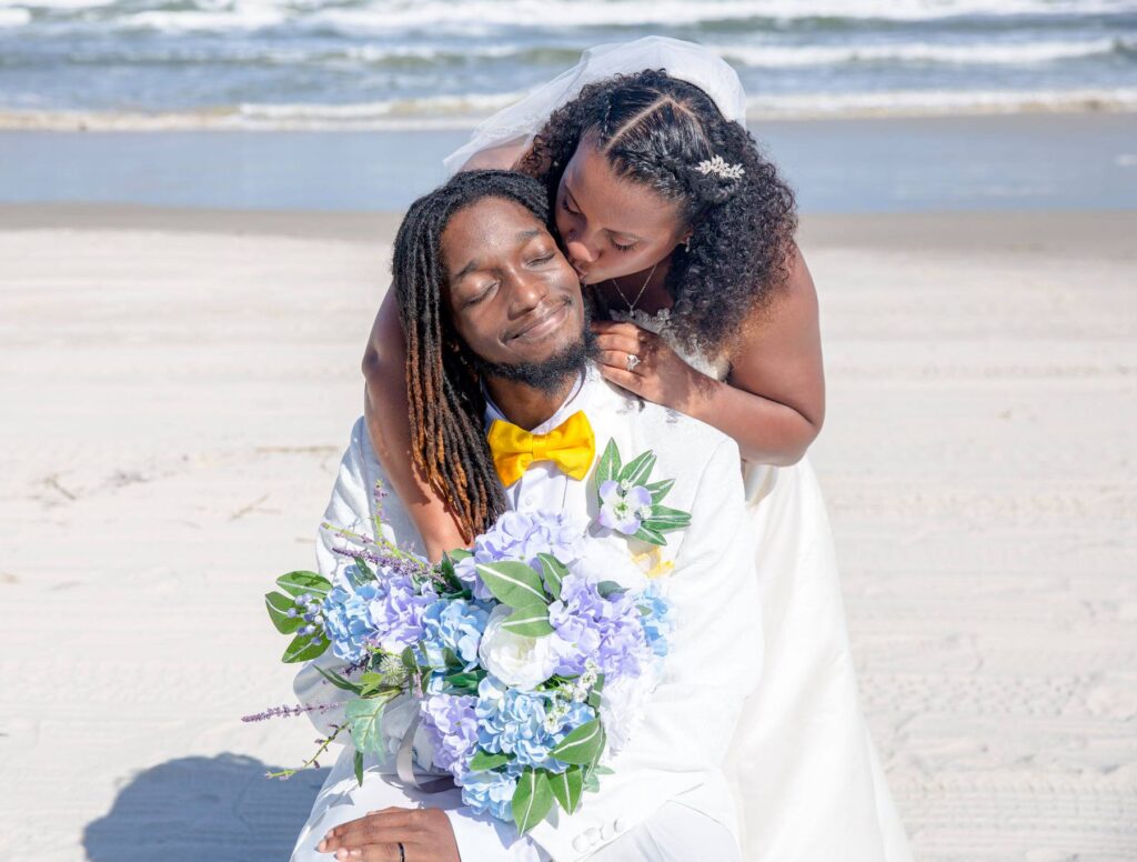 groom in white suit and yellow bow tie sitting on beach with bride leaning down behind him to kiss him on the cheek