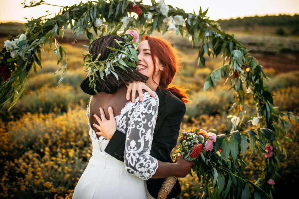 bride in white dress and floral crown hugging bride in black jacket in front of floral arch in a field