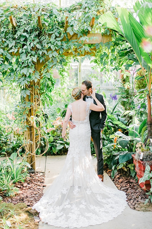 A bride and groom under a garden arch. The bride's back is to the camera and the groom is kissing her cheek.