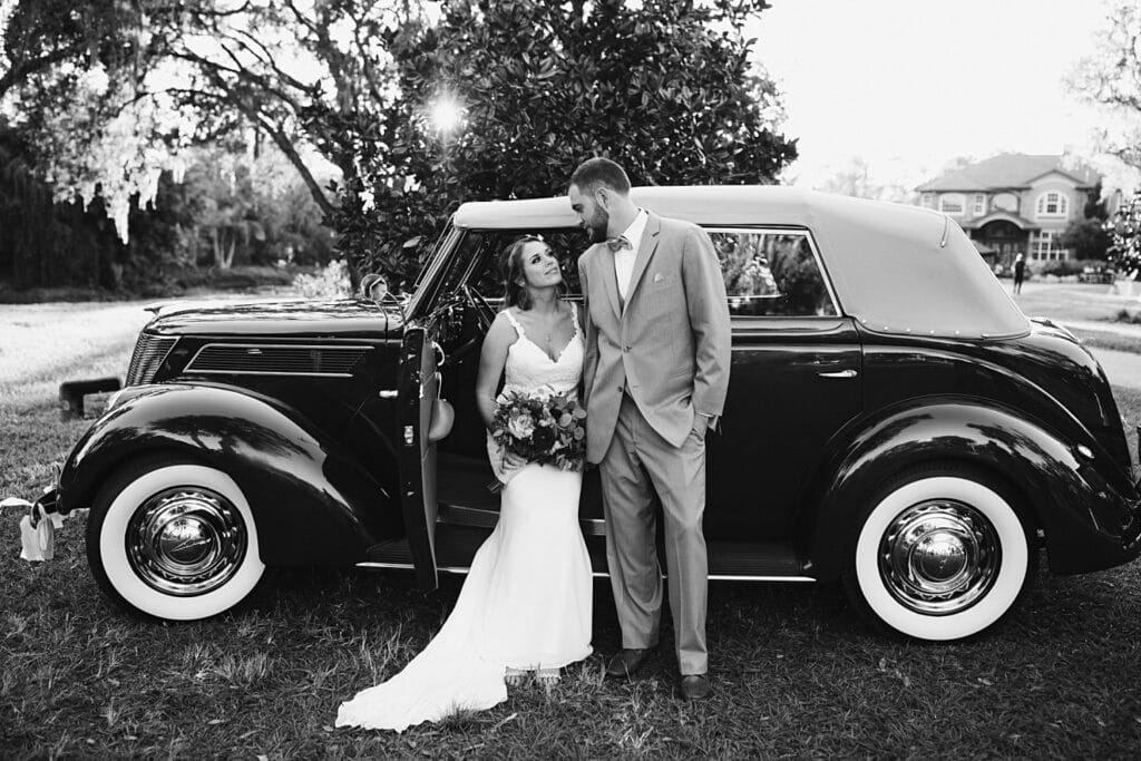 A bride and groom in front of an old style car. The photo is in black and white and the bride and groom are looking at each other happily.