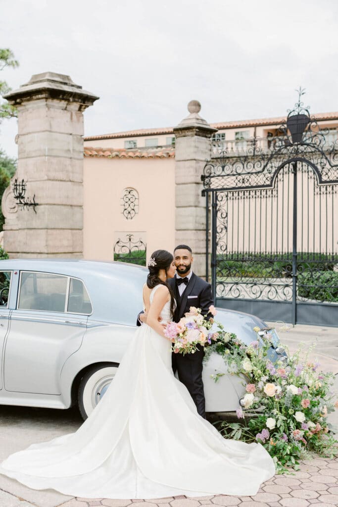 Bride and groom standing in front of an old style car. There are floral arrangements behind the car.