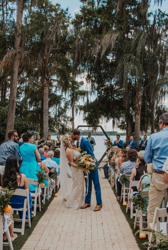 Bride and Groom kissing in aisle with groom in blue suit and lake in distance