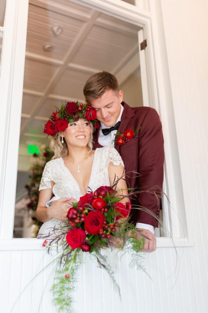 Christmas in July SS 122 - Christmas Themed Wedding Inspiration - Inspiration Shoot groom hugging the bride from behind while the bride smiles and is holding her red and green bouquet.