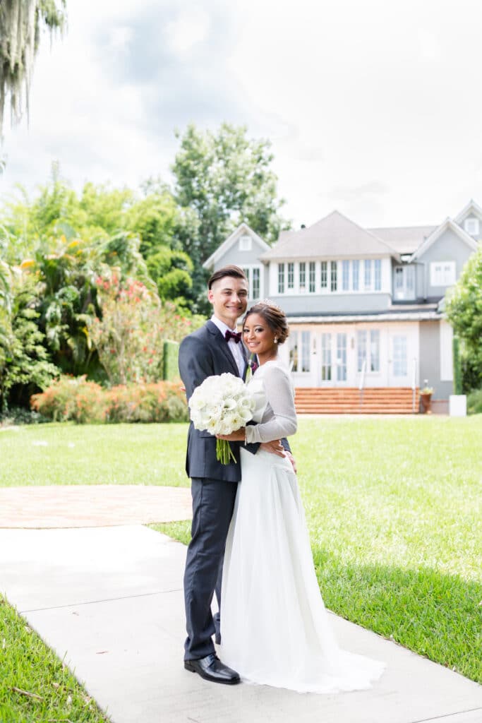 Christmas in July SS 164 - Christmas Themed Wedding Inspiration - Inspiration Shoot Bride and groom standing outside of their wedding venue. They are facing the camera and the background has a large chic building and a big garden.