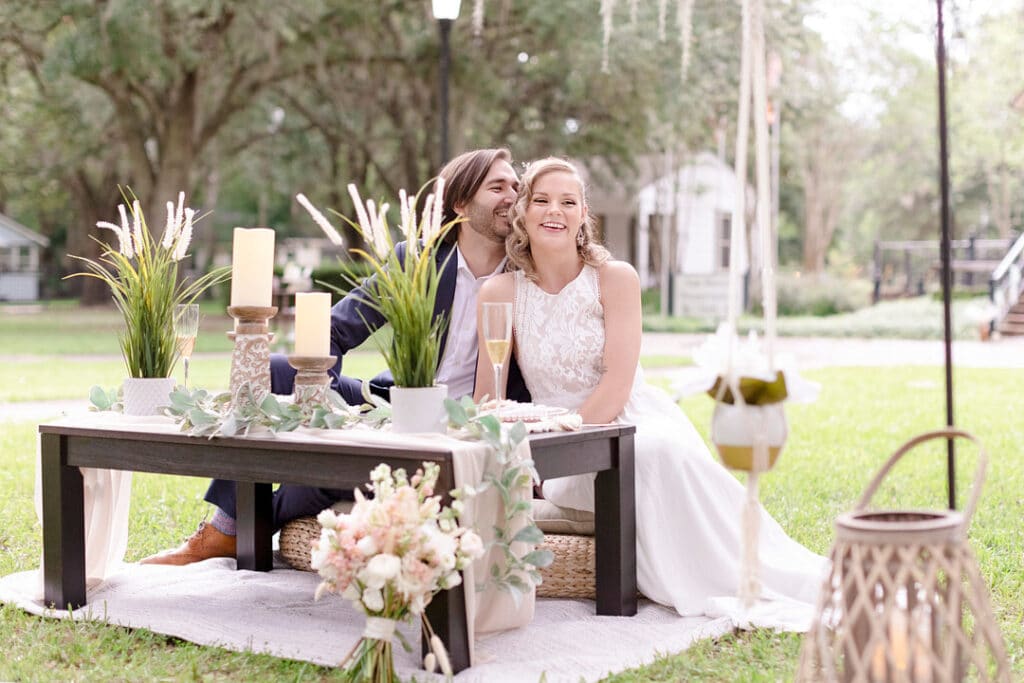 A bride and groom sit on rattan pillows at a small table with candles, glasses of champagne and green ivy. The setting is outdoors.