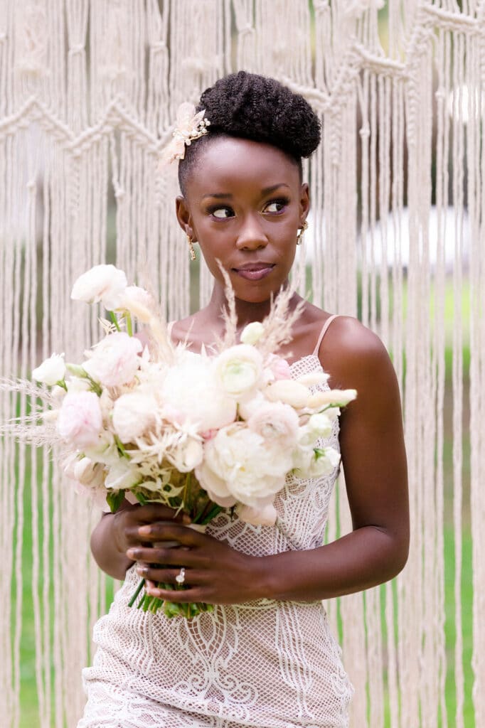 bride looking to the right and holding her bouquet with a macrame backdrop in the background for the picnic wedding inspiration shoot