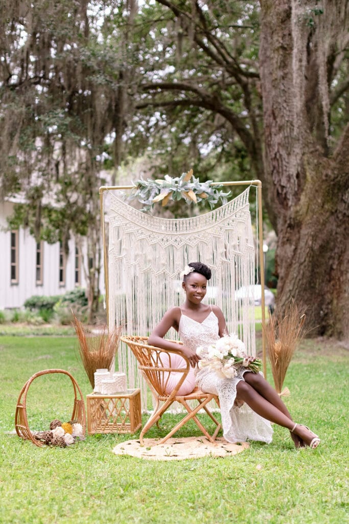 Bride sitting in a wicker chair holding a boutquet of white flowers. She is in front of a macrame tapestry and is sitting outside.