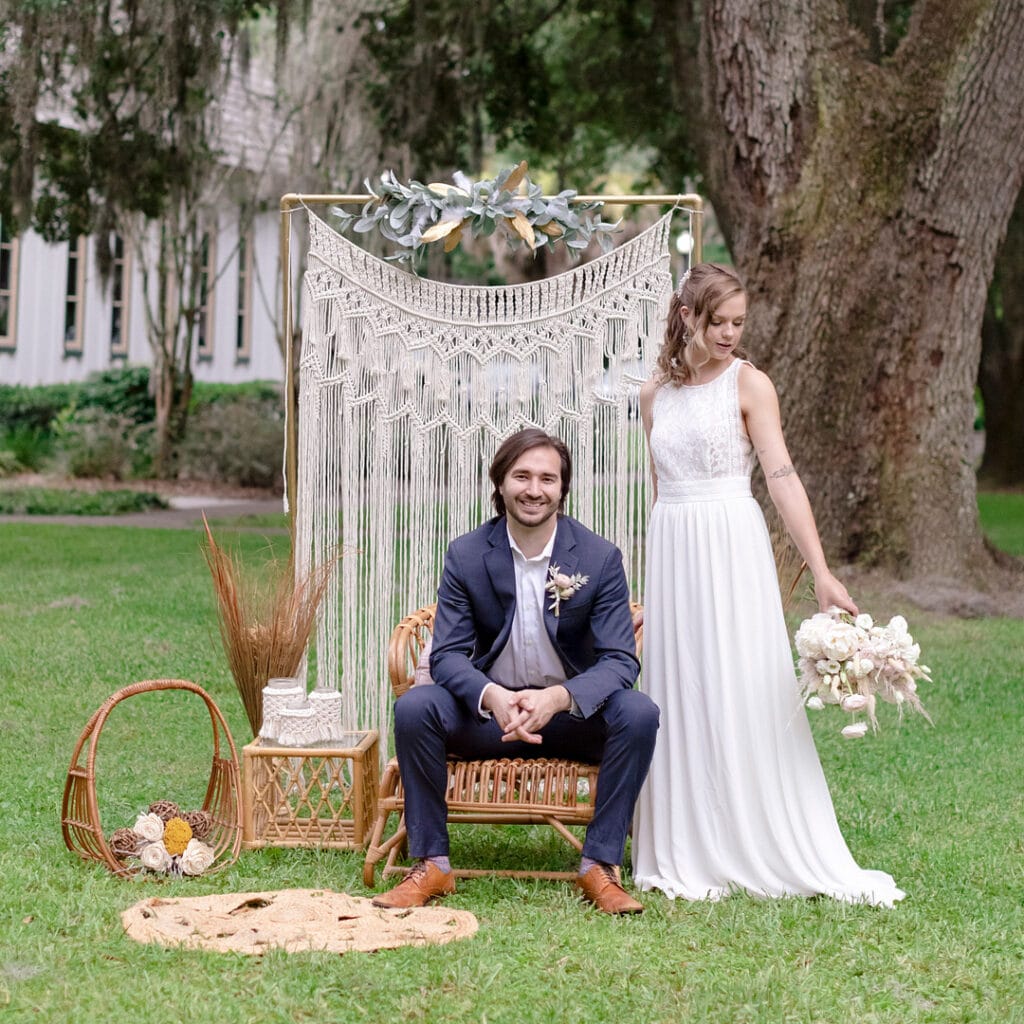 Bride and groom in front of a macrame tapestry. They are outside in front of a tree and the groom is smiling
