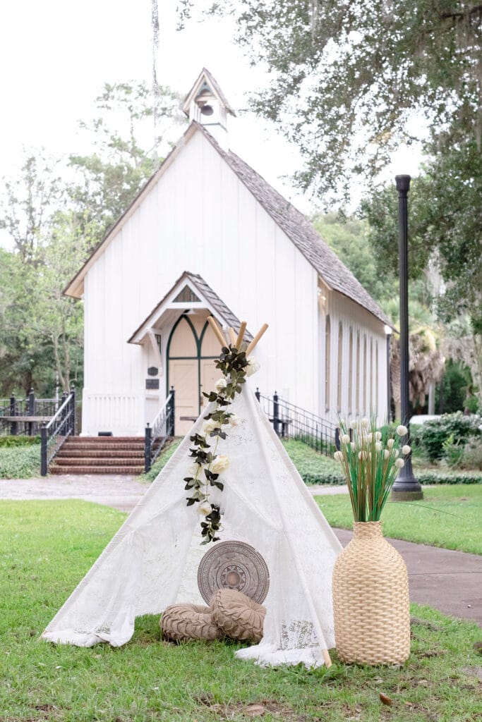 Boho chic lace tent with pillows, a vase, and white flowers. They are outside in front of a church.