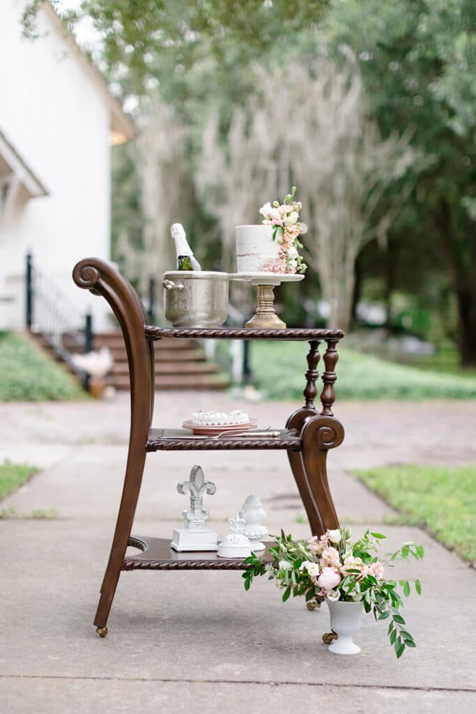 Wooden bar cart with chic decor. The top of the cart has a bottle of champagne and a simple, yet beautiful, white wedding cake.