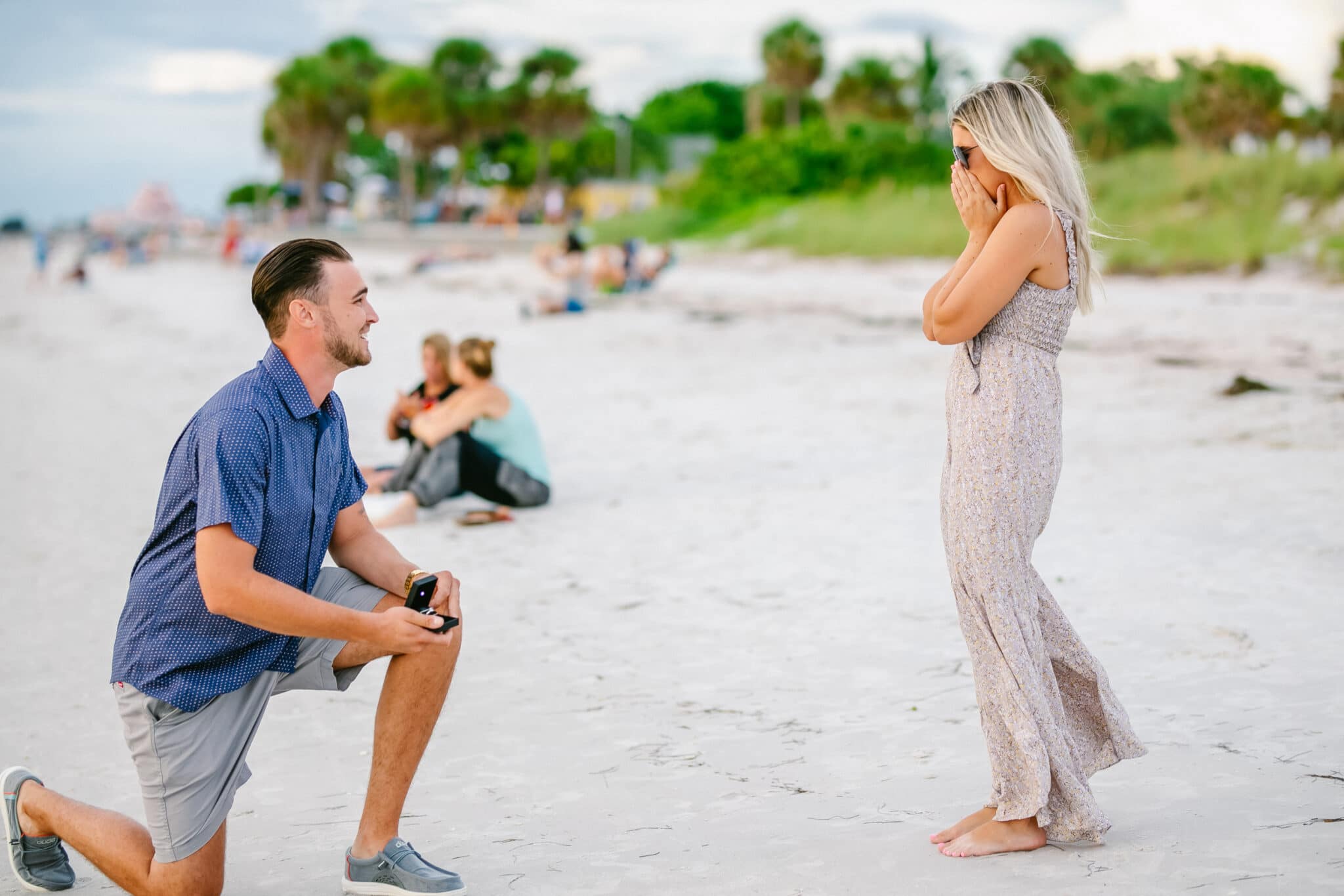 groom to be on one knee proposing to her bride to be during their sunset on the beach marriage proposal