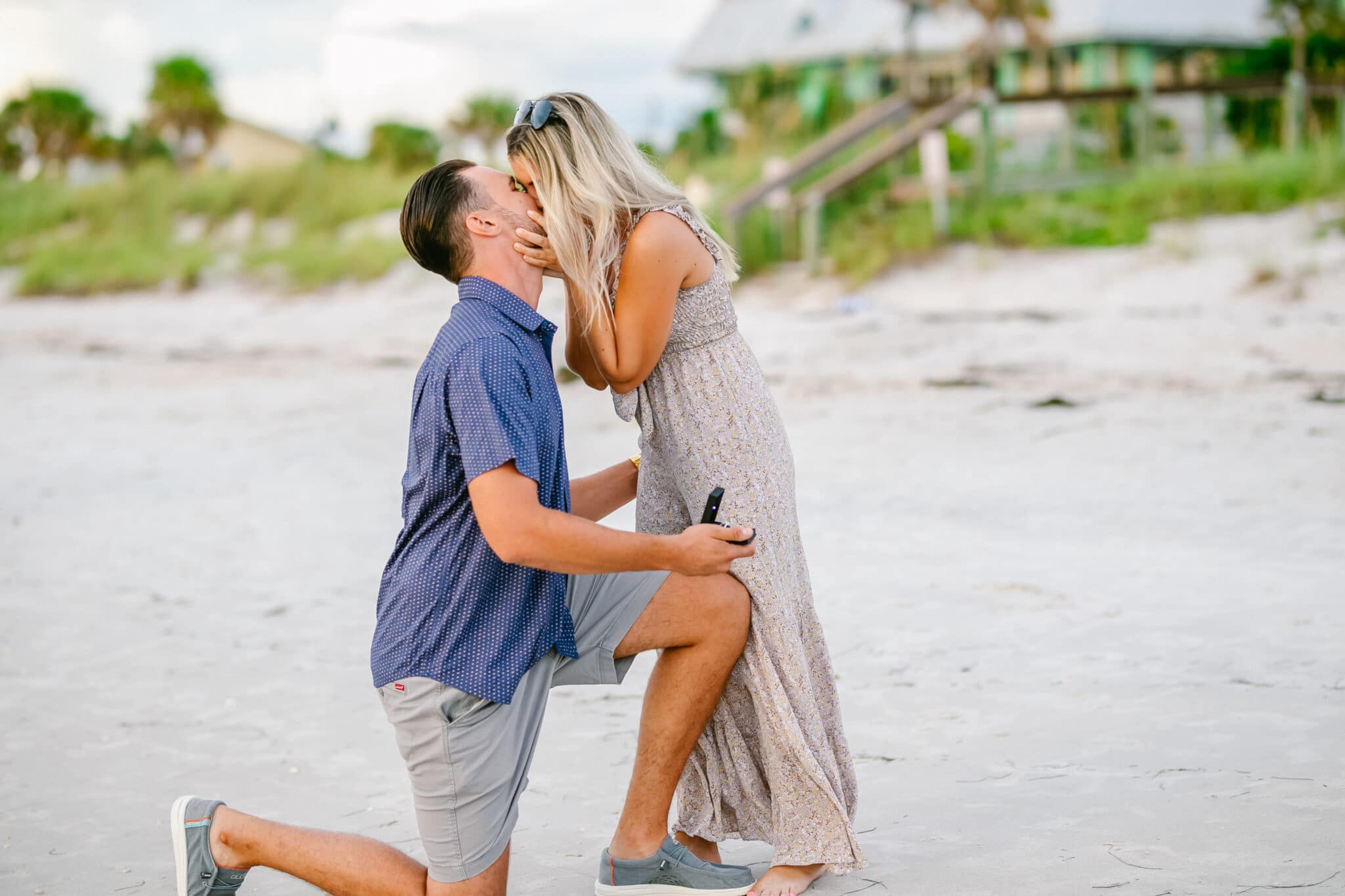 bride and groom to be kissing on the beach while the groom is still on one knee after proposing.