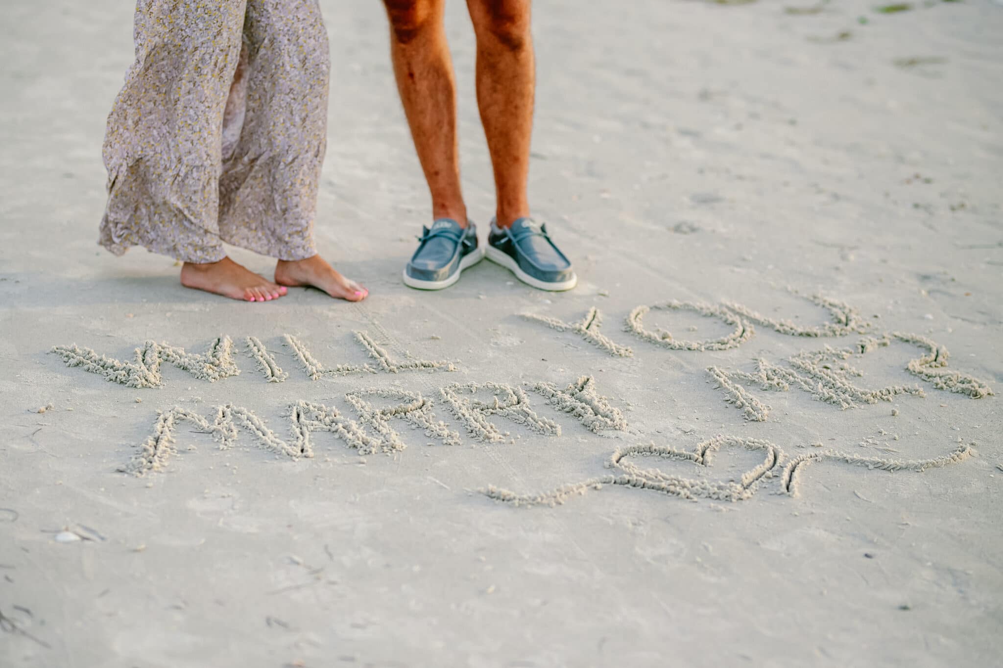 newly engaged couple standing above the words "will you marry me" written in the sand.