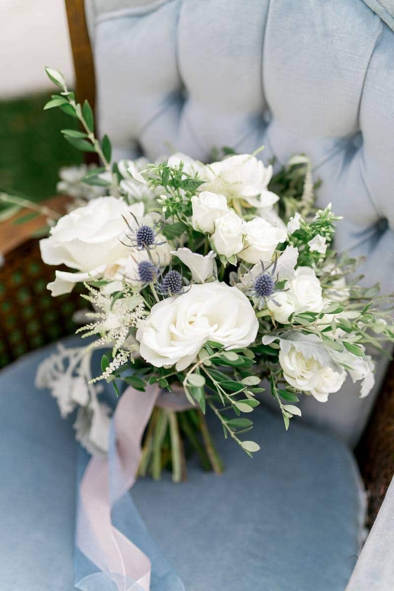 bouquet at wedding sits on velvet blue chair with long ribbon around it and white flowers with unique greenery and wild flowers