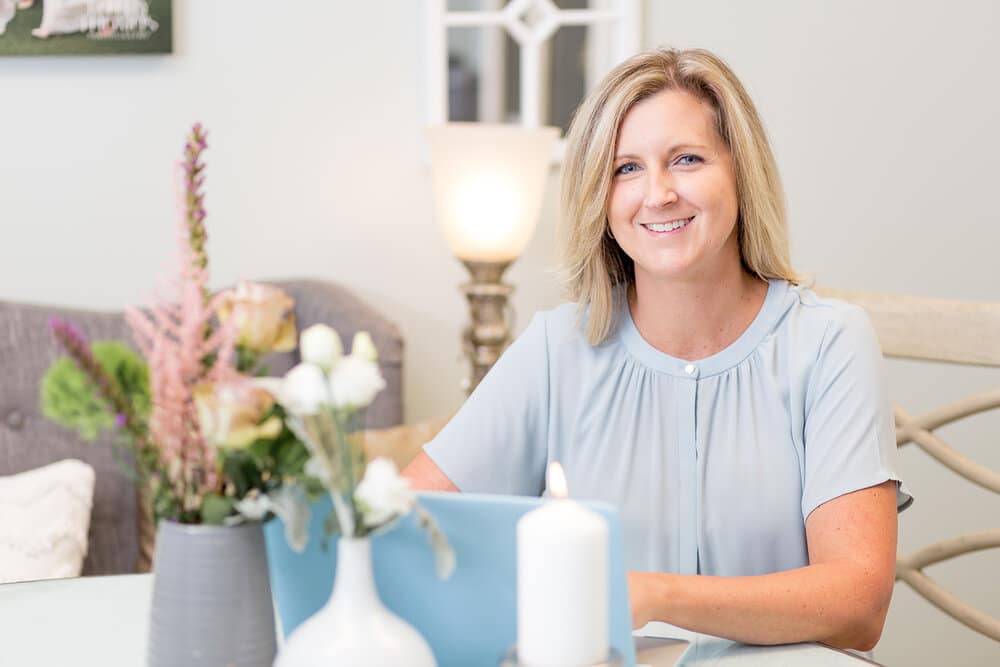 woman in light blue shirt sits at desk with laptop in front of her and flowers in a vase on table along with tall candle