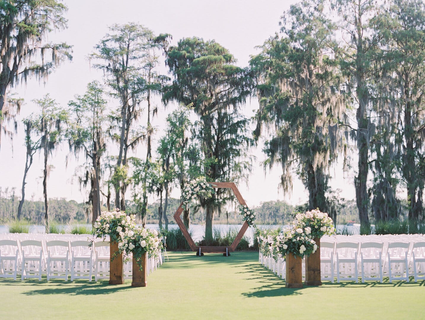 wedding ceremony setup in front of body of water with end of aisle floral arrangements and hexagon wooden arch at the altar with white chairs for the guests