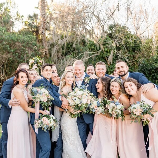 wedding part with groomsmen in blue and bridesmaids in light pink surrounding bride and group with the bridesmaids holding bouquets