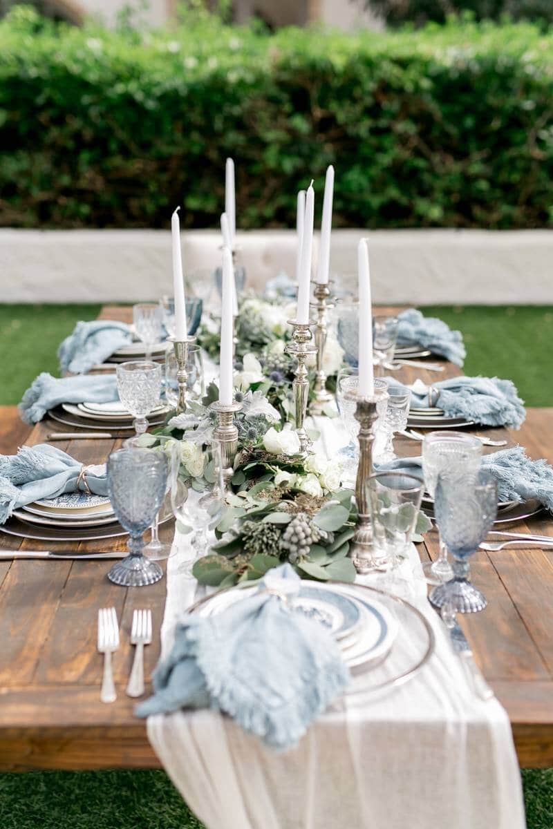 tablescape from wedding table with charges, place settings, water goblets, tall candlesticks with holders and greenery down the middle of the long wood table