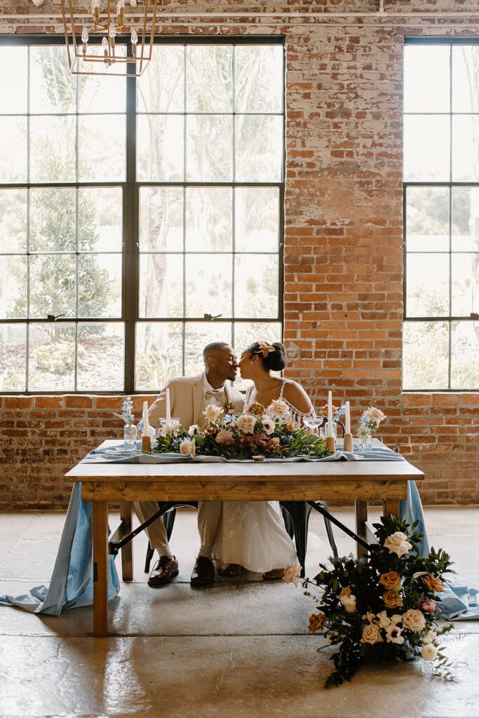 Summer Urban Loft Wedding Inspiration Grace Jicha Venue At The Block Wedding BrideandGroom 48 websize low - Urban Loft Wedding Inspiration - Styled Shoot bride and groom leaning in for a kiss while sitting at their reception table for the urban loft wedding inspiration post.