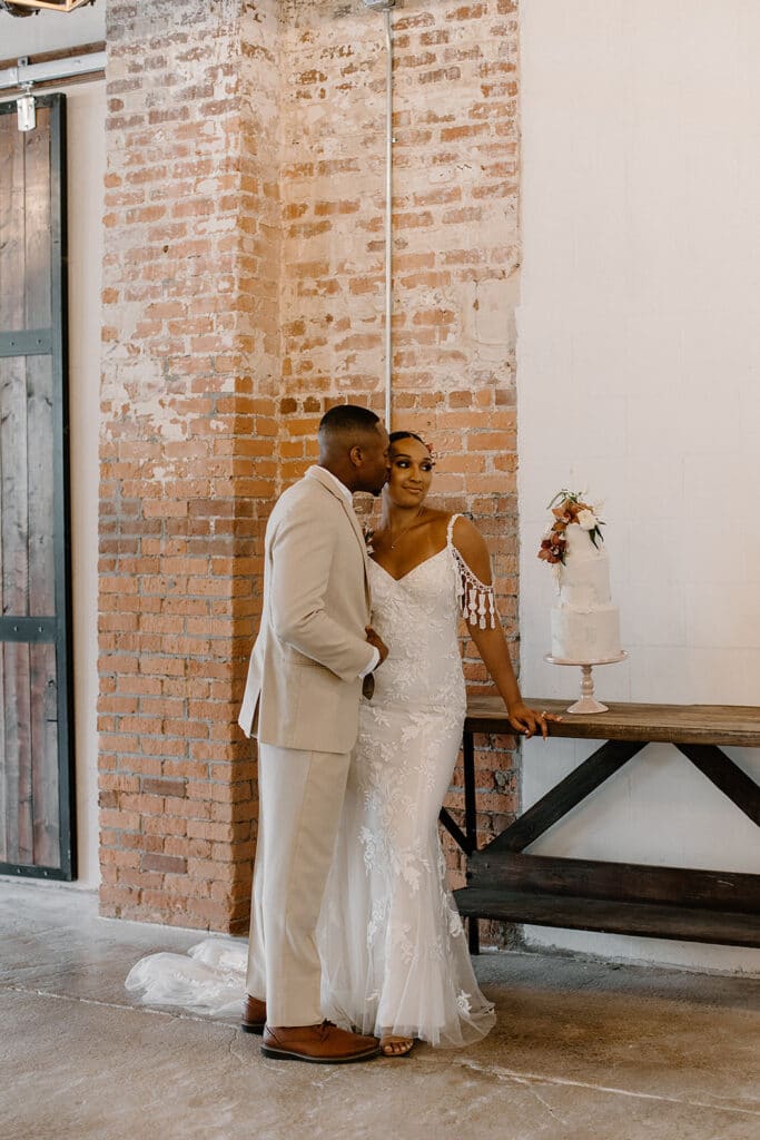 Summer Urban Loft Wedding Inspiration Grace Jicha Venue At The Block Wedding Cake 13 websize low - Urban Loft Wedding Inspiration - Styled Shoot groom kissing the bride on the cheek next to their three tier cake.