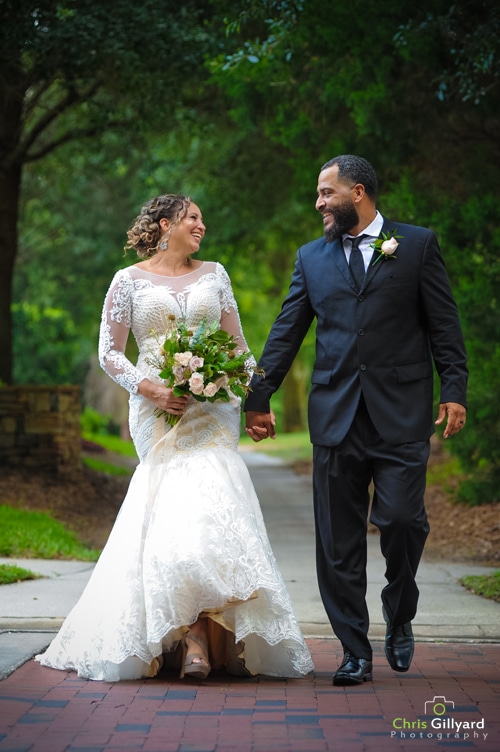 Bride and groom walking under trees. They are looking at each other and smiling. The bride is holding an arrangement of flowers.