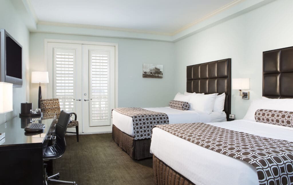 hotel room with two beds, workspace, light blue walls, and double doors leading out to balcony
