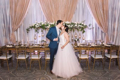 Bride and groom kissing in front of their bridal table. They are holding glasses of champagne.