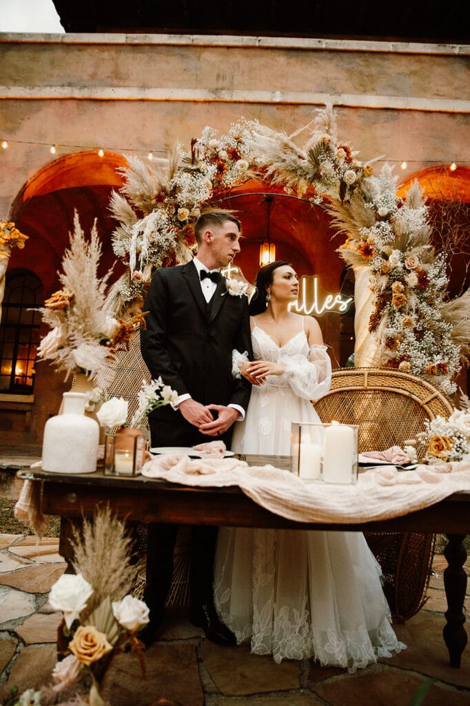 A bride and groom looking off into the distance. They are standing behind a rustic wooding table adorned with candles and flowers.