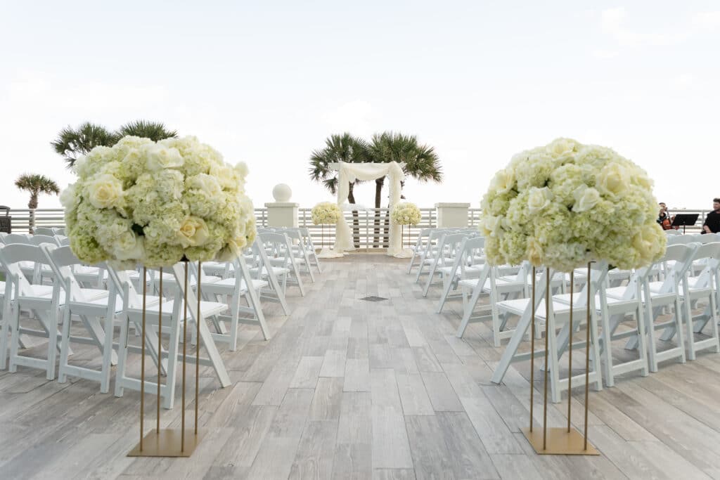 wedding setting with white chairs, tall white flower arrangement and arbor in distance