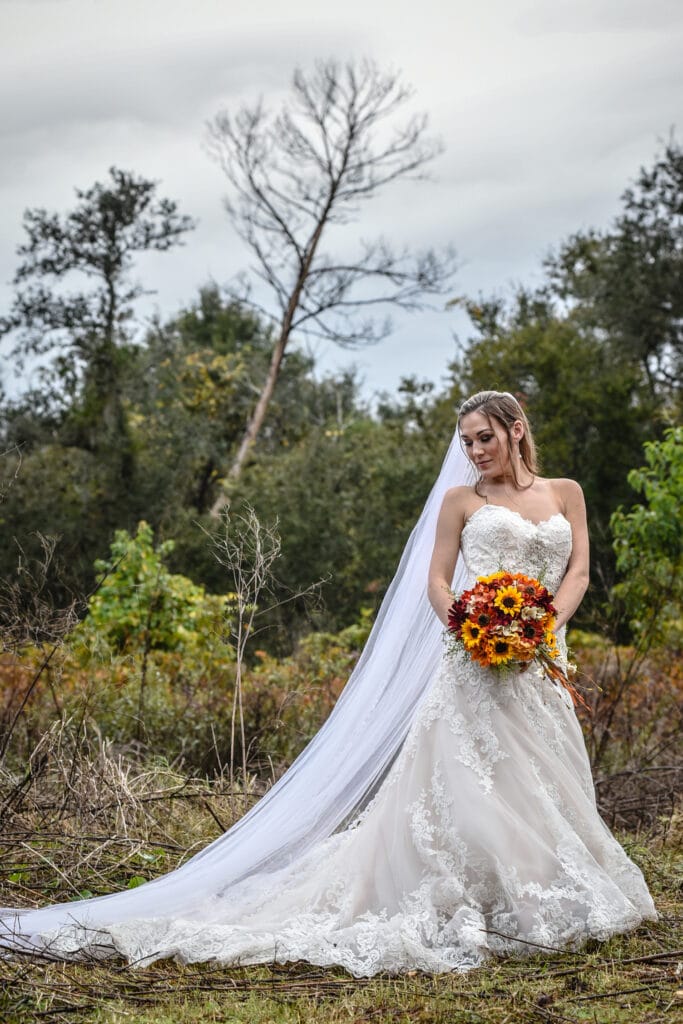 Bride with a sunflower and fall bouquet. She is looking at the ground and is in front of the trees