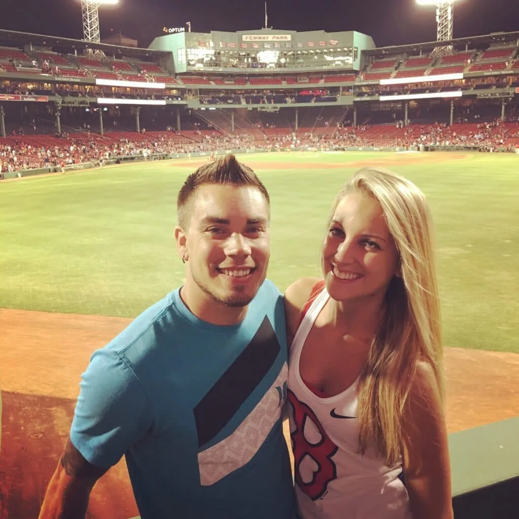 newly engaged couple smiling in front of a baseball field.
