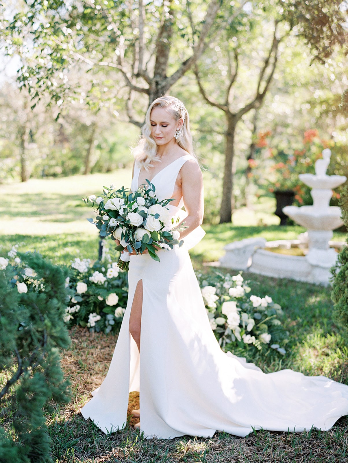 Bride in a garden holding a bouquet of white flowers. She is standing in front of a decorative fountain