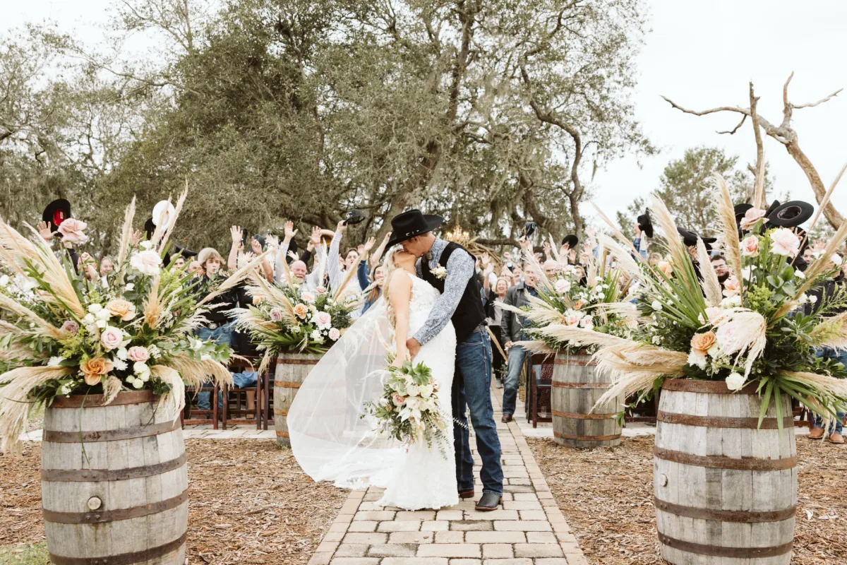 Bride and groom kissing during a Western wedding ceremony surrounded by pampas grass, blush roses, white flowers, and lush greenery.