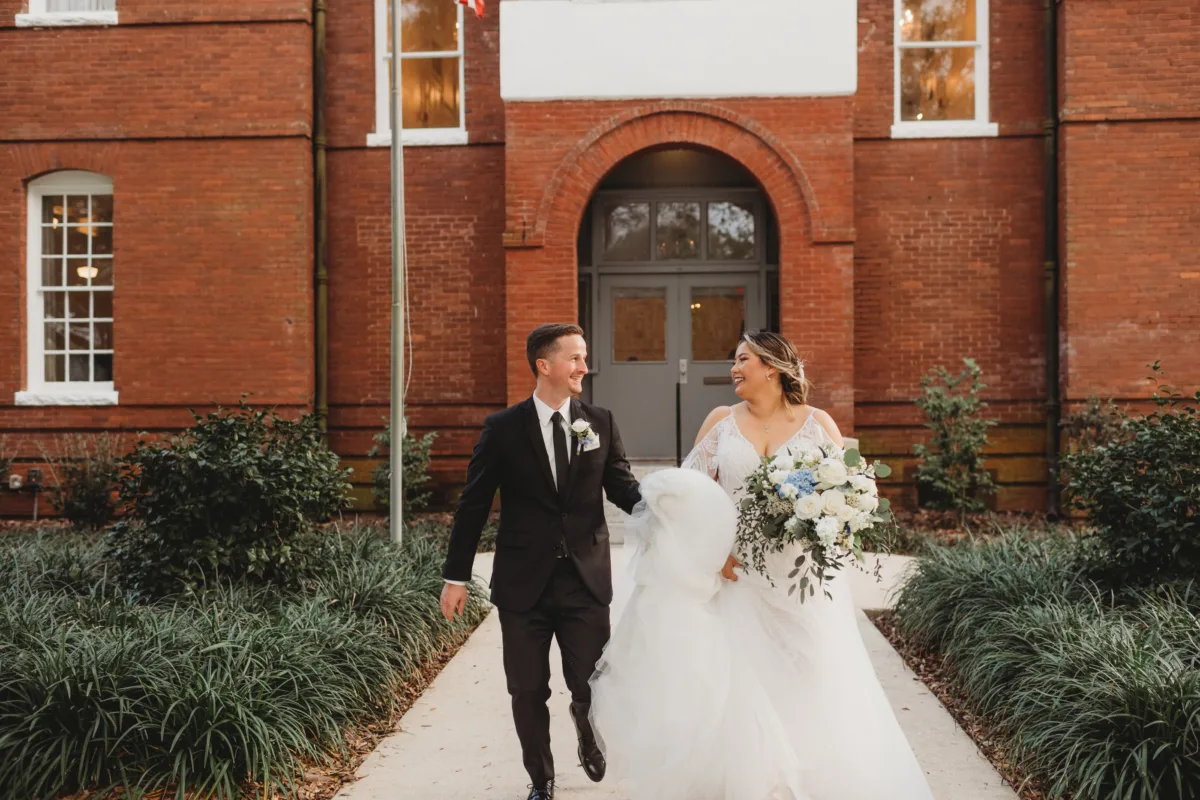 Bride and groom walking outside a historic brick wedding venue with an elegant white and blue bridal bouquet.