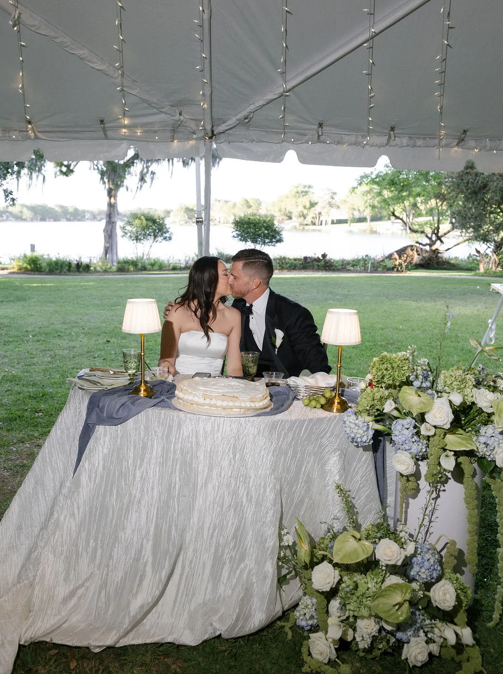 Bride and groom kissing at an elegant lakeside wedding reception surrounded by white and blue floral arrangements.