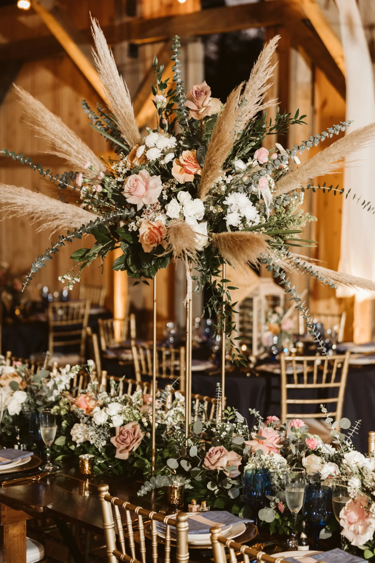 Luxury barn wedding reception featuring a tall floral centerpiece with pampas grass, blush roses, white flowers, and eucalyptus.