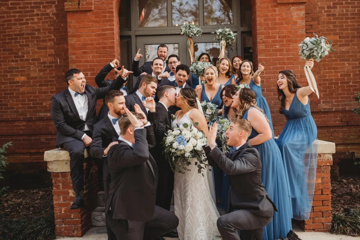 Joyful bride and wedding party posing outside a brick wedding venue with white and blue bouquets.
