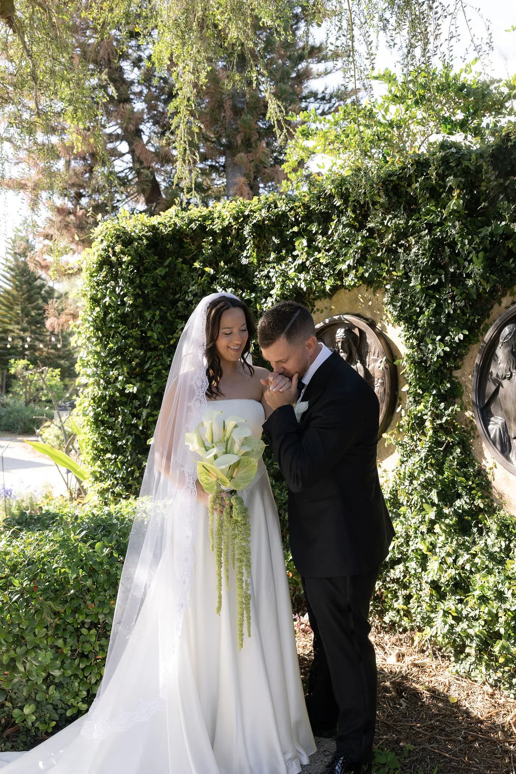 Bride and groom sharing a romantic moment outdoors with an elegant white calla lily bouquet and cascading green flowers.