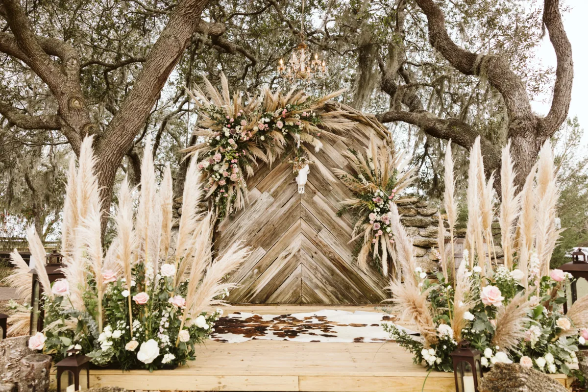 Luxury outdoor wedding ceremony with a rustic wood backdrop, pampas grass, blush roses, and lush floral arrangements beneath oak trees.