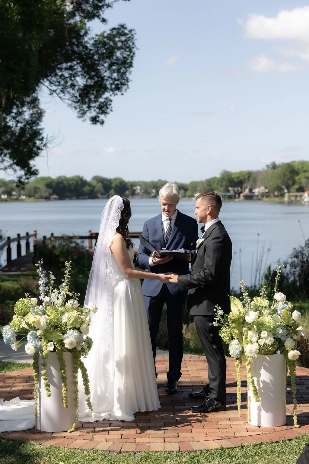 Bride and groom exchanging vows beside a lake surrounded by elegant white, blue, and green wedding flowers.