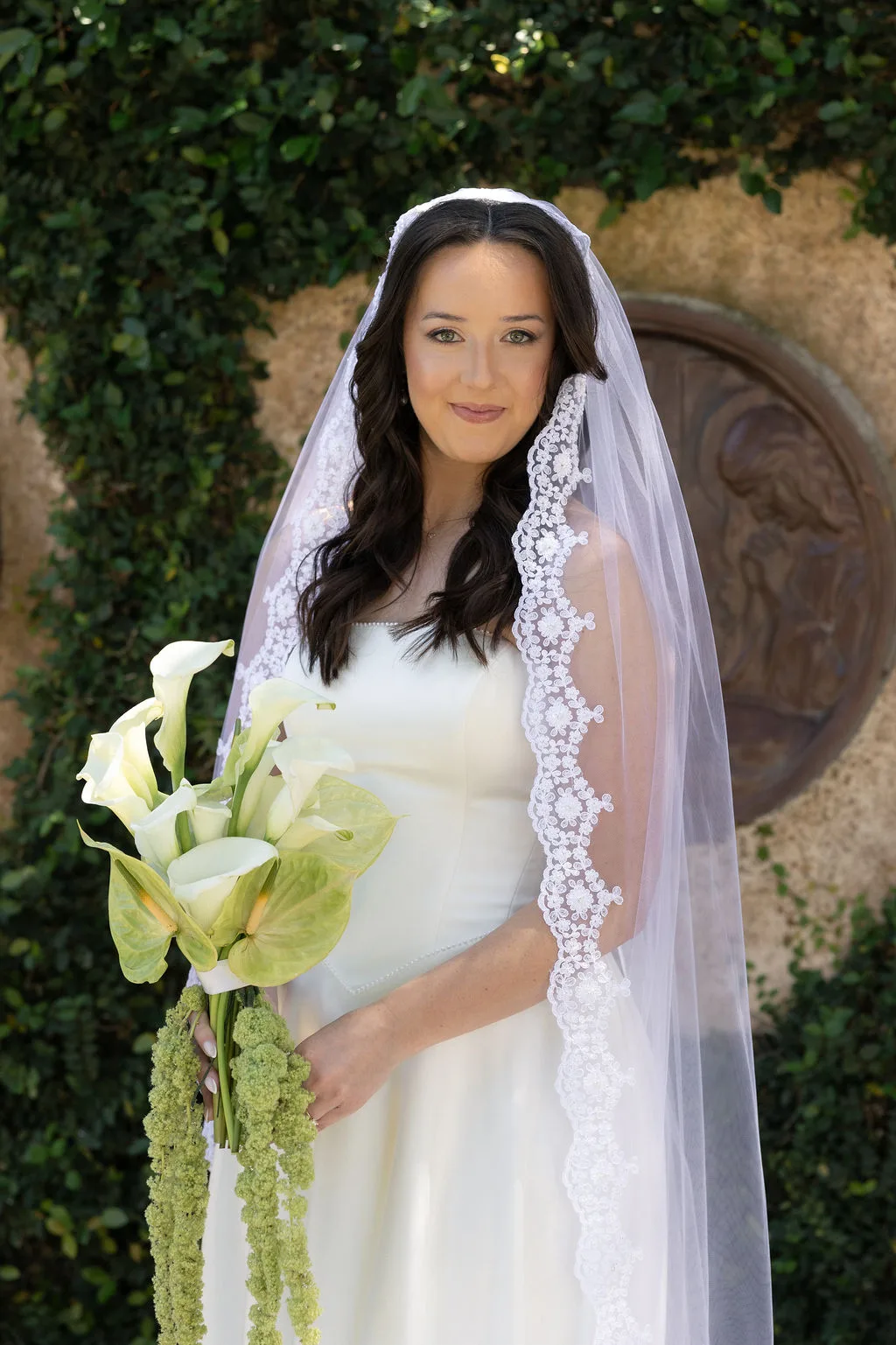 Bride in an elegant lace-edge veil holding a sculptural white calla lily bouquet with cascading green flowers.