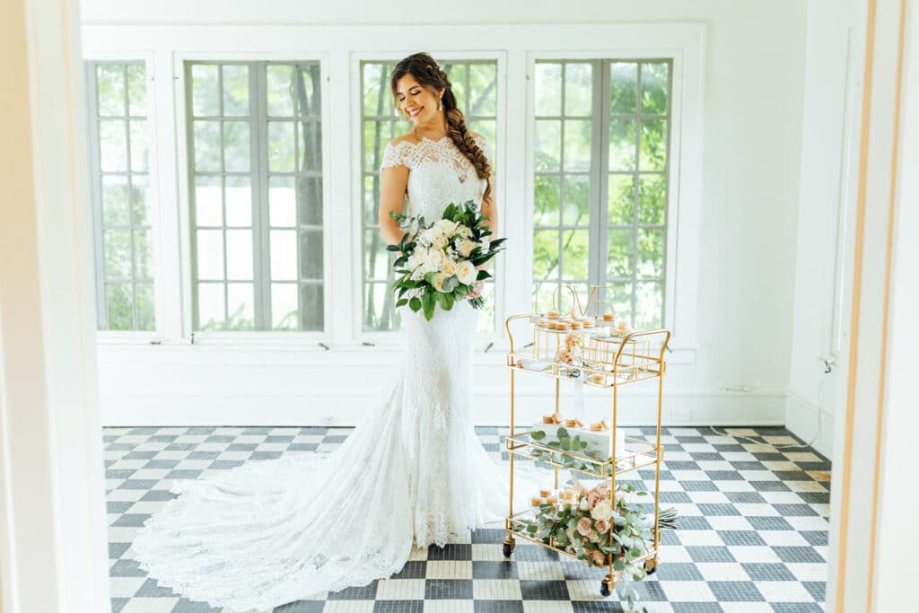 bride holding white bouquet and standing next to gold cart in a white room with black and white tile floor