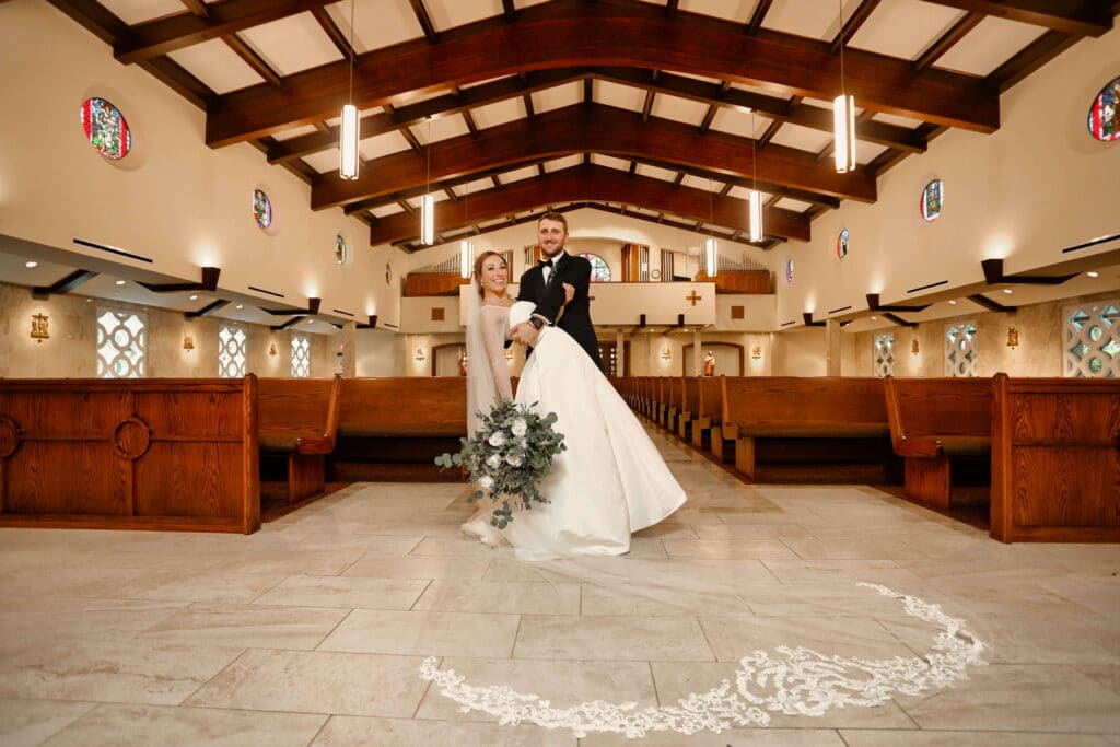 Bride and groom in a church. The bride's veil is spread out in front of them and the groom is leaning the bride backwards.