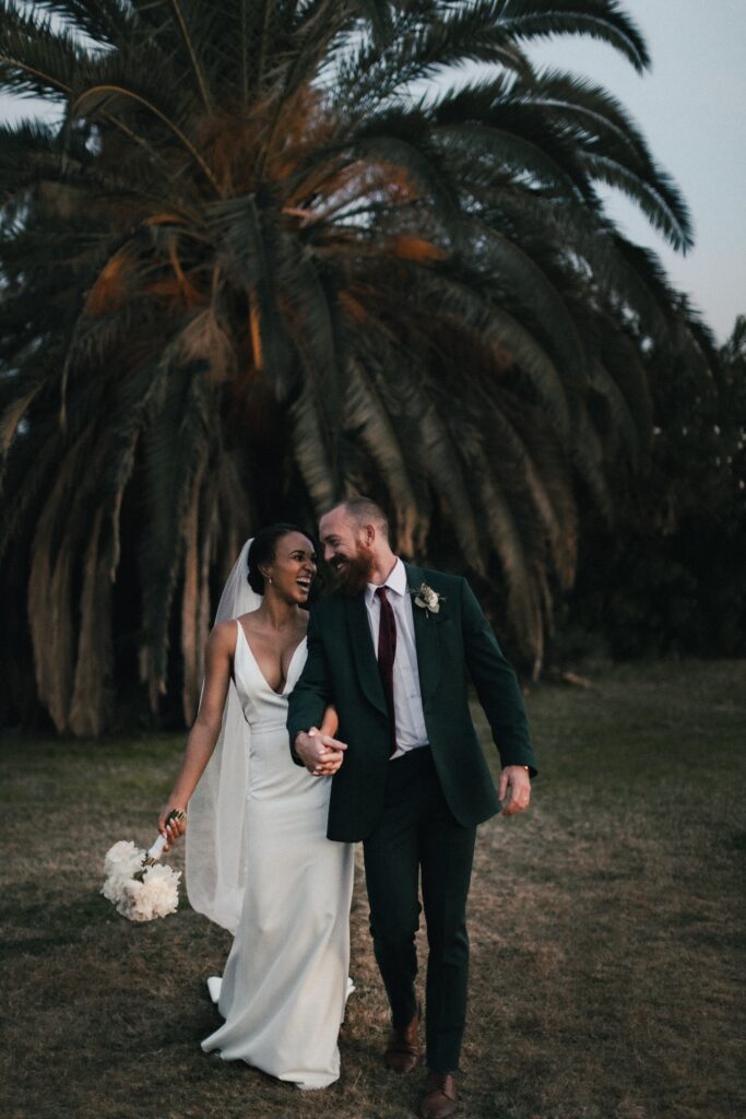 bride and groom walking arm in arm and looking at each other with palm tress in the background
