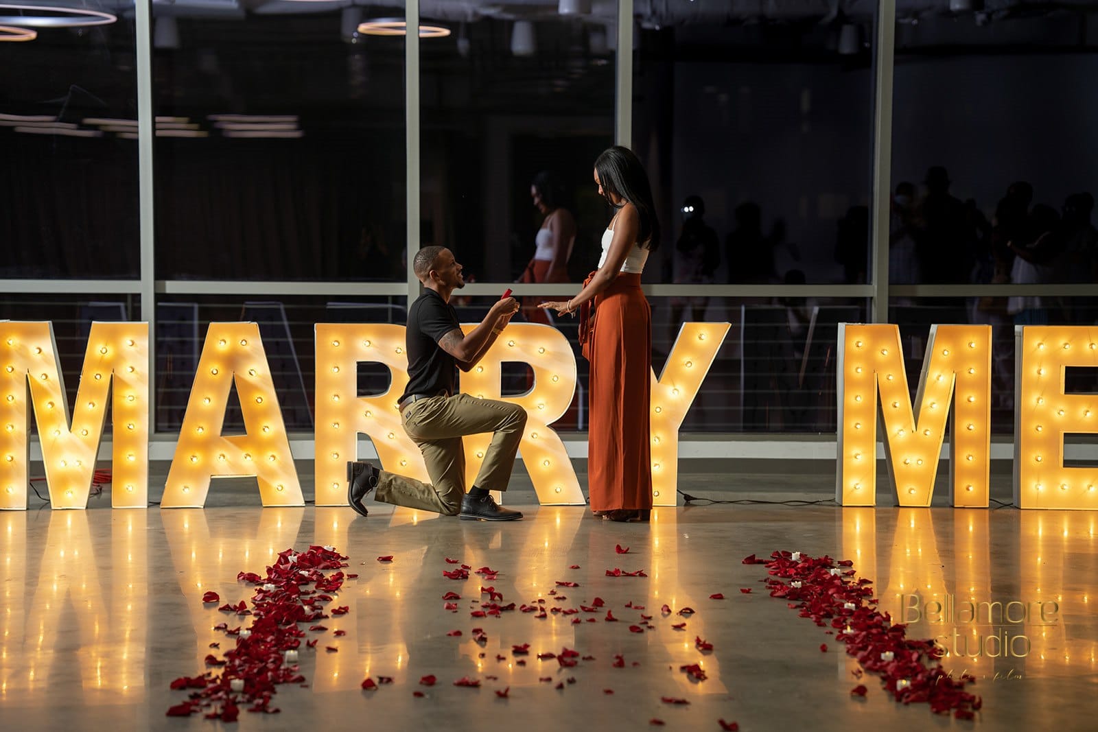 groom to be on one knee proposing to his girlfriend in front of a marry me sign surrounded by rose petals during their downtown orlando marriage proposal.