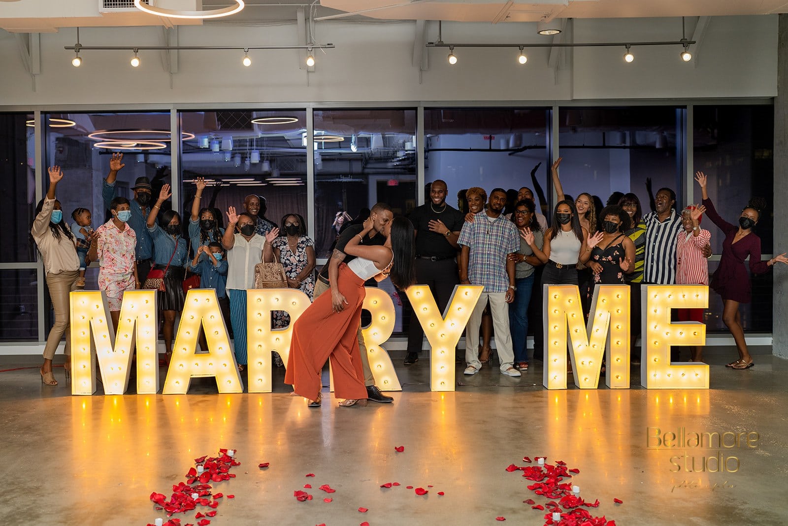 newly engagec couple kissing in front of the marry me sign after their downtown orlando marriage proposal with their friends and family behind the sign cheering.