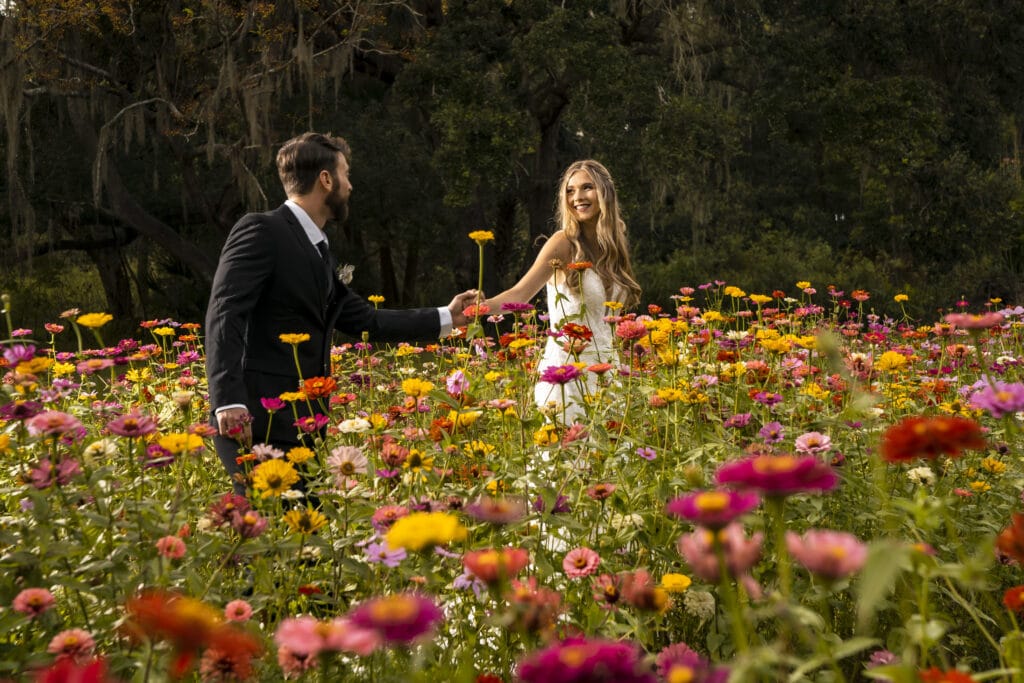 bride and groom holding hands and looking at each other while walking through field of flowers