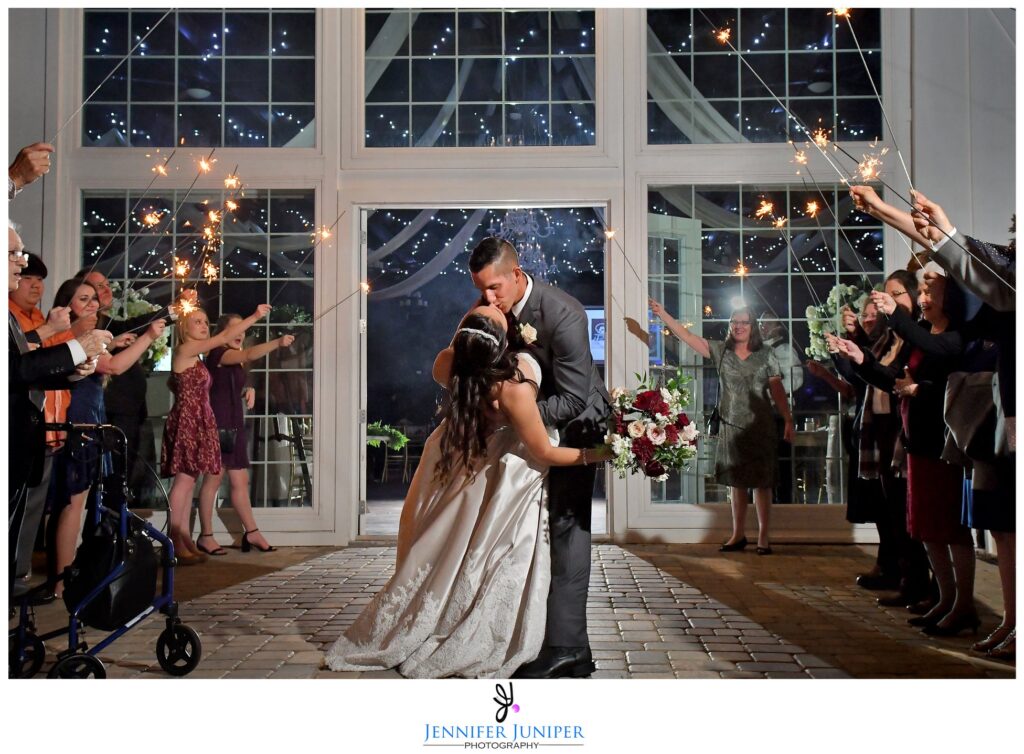 bride and groom kissing in front of wall with large windows while wedding guests hold sparklers
