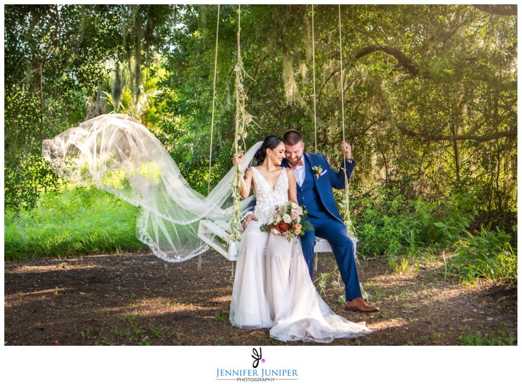 bride and groom sitting together on wooden swing with her veil flowing in the wind